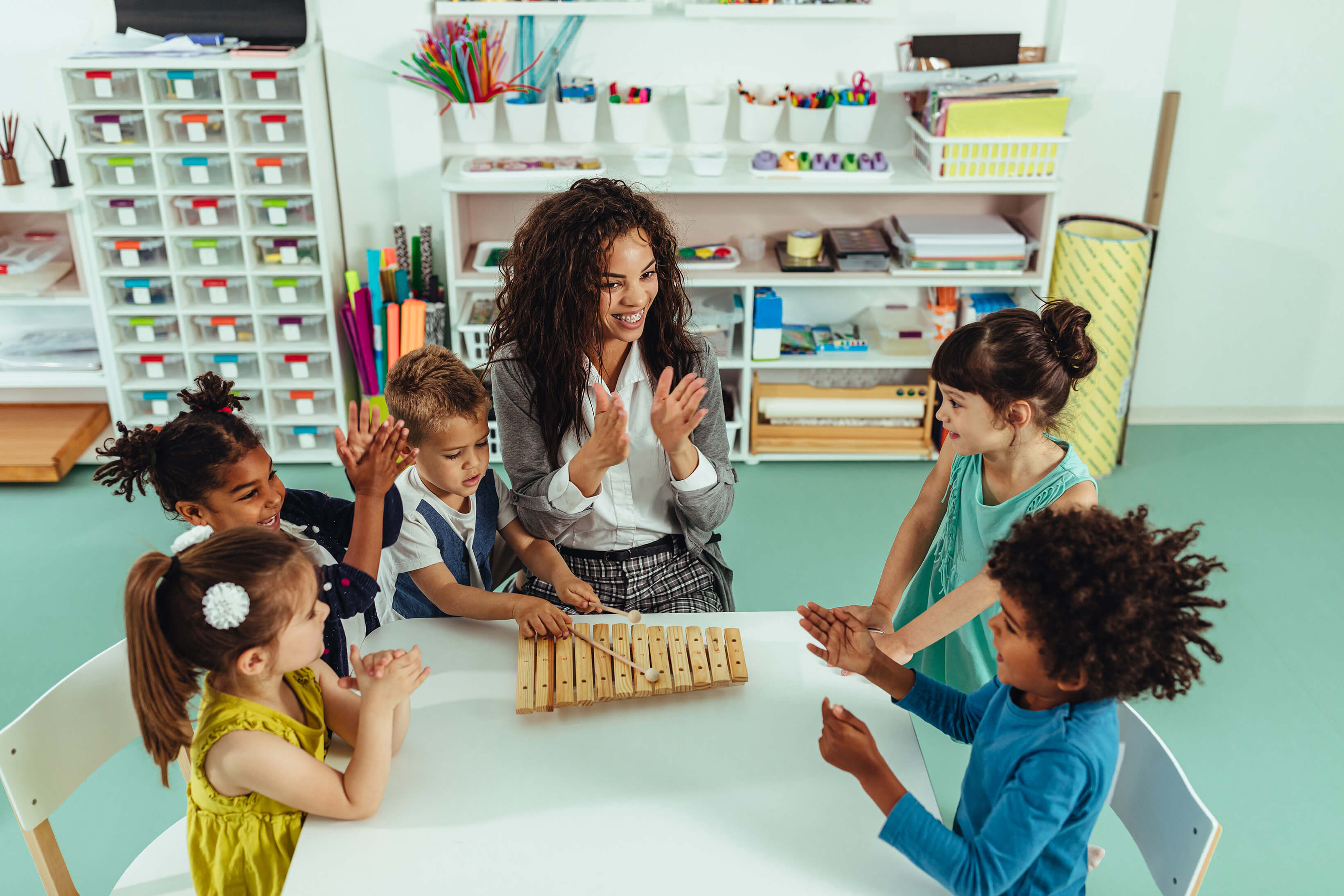 A teacher and group of students sitting at a table, playing with a xylophone.